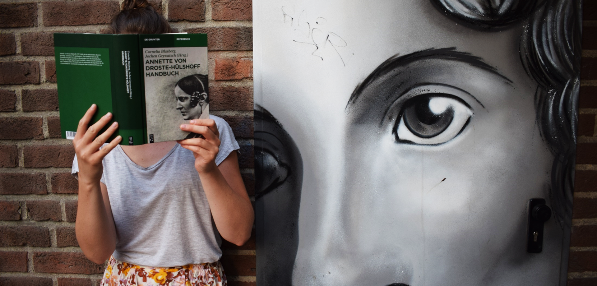 Frau mit Droste-Handbuch neben einem Graffito der Autorin Annette von Droste-Hülshoff (erstellt von Lackaffen) in der Schillerstraße in Münster.